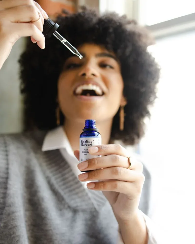 Smiling woman with a full natural afro holding a scalp serum/ hair oil bottle and glass dropper.