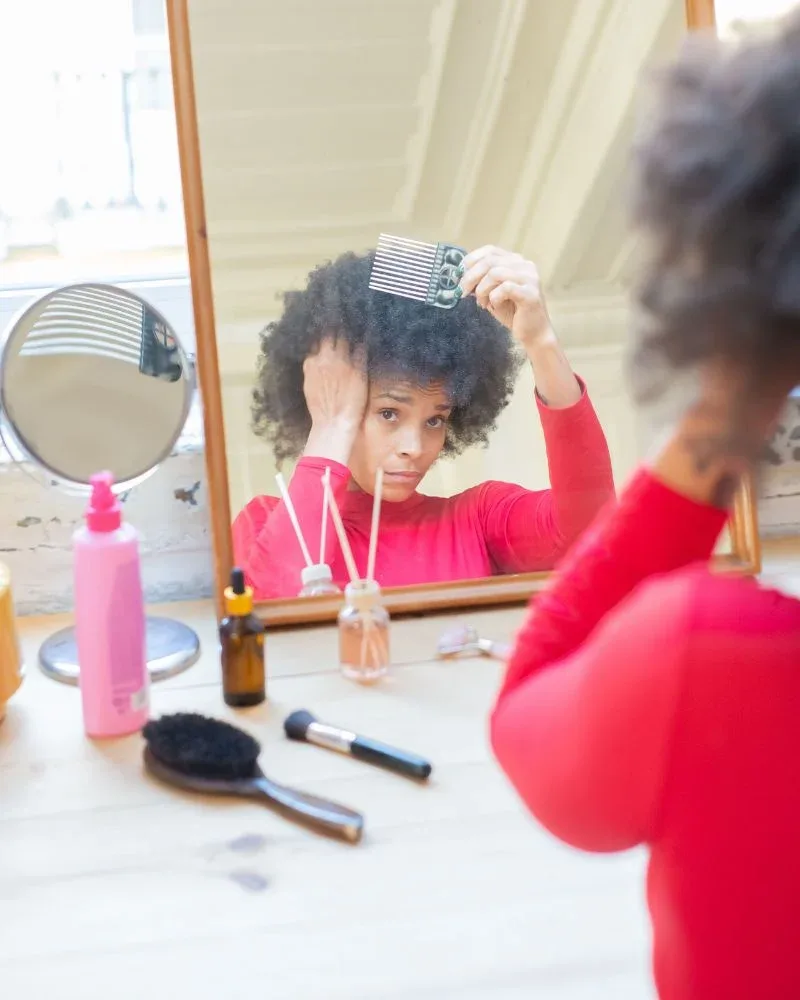 Woman detangling fine 4C hair with an afro pick while looking in a mirror